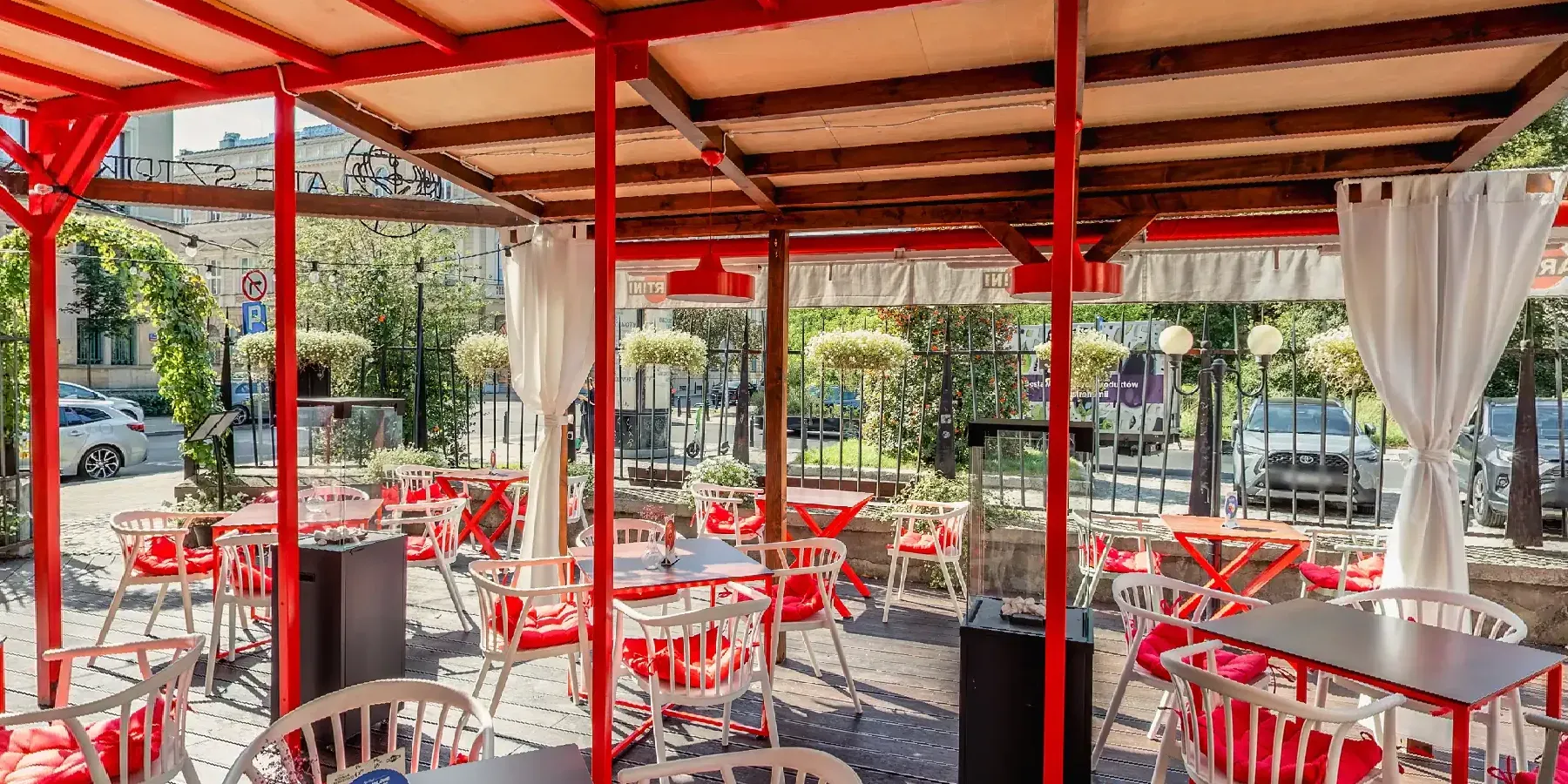 Taras restauracji Ale Sztuka w Warszawie: czerwone zadaszenie, białe krzesła z czerwonymi poduszkami i ogród za ogrodzeniem. Kadr ze spaceru 360°.