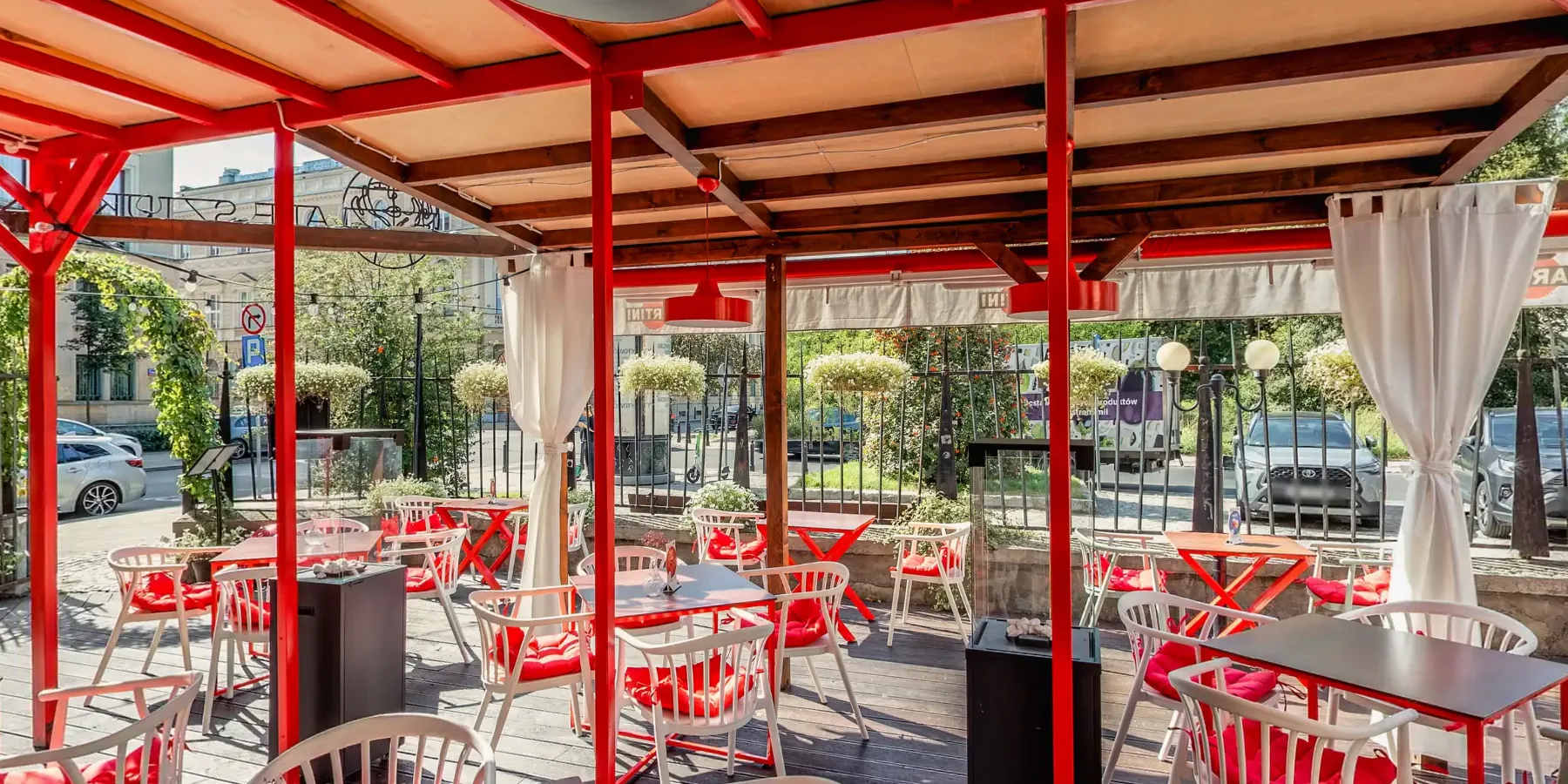 Taras restauracji Ale Sztuka w Warszawie: czerwone zadaszenie, białe krzesła z czerwonymi poduszkami i ogród za ogrodzeniem. Kadr ze spaceru 360°.